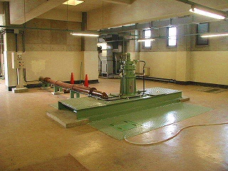 茶色がかった広い床とコンクリートの壁を持つ広い空間の中央に、緑色の台座に設置された工業用ポンプまたはモーターと太い配管設備を写した写真