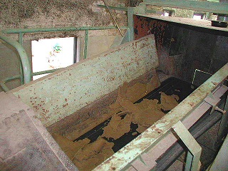 汚泥処理場内にある、表面が錆びて塗装が剥がれた大きな金属製のホッパーが、乾燥した茶色い汚泥や粉体のような物質で満たされている様子を、上部からやや斜めに見下ろした写真