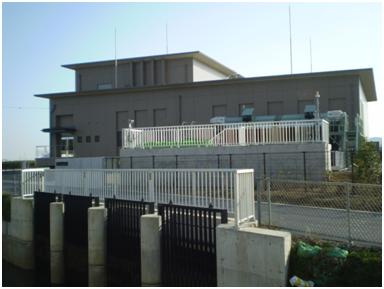 淡い茶色とグレーの平らな屋根を持つ二階建ての建屋が、複数の白と黒の柵やフェンスで厳重に囲まれ、その手前にはコンクリートの構造物と水面が見えるポンプ場または水処理施設の外観写