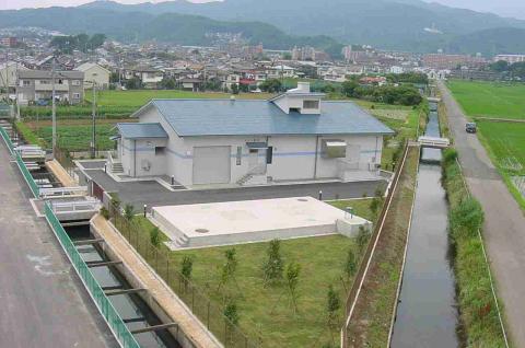 淡いグレーの外壁と青い屋根を持つ近代的な建物が、住宅地の近くの水路と田畑に囲まれた3角形の土地に建っている、鶴巻中継ポンプ場の写真