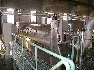 水処理施設内部で、頑丈な金属製のデッキの上に設置された、覗き窓のような楕円形のハッチが複数ついた巨大な円筒形のスクリュープレス脱水機を捉えた写真