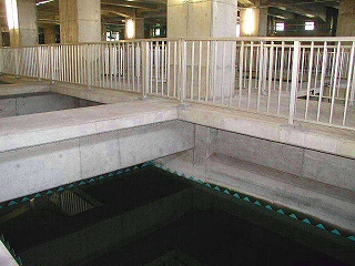コンクリートの柱が並ぶ広大な屋内施設で、手すりのついた通路の下に黒い液体が入った大きな水槽が並んでいる写真
