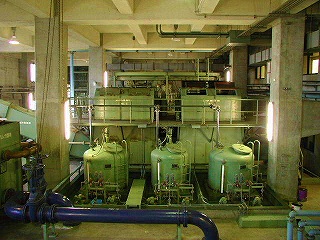 照明に照らされた水処理施設で、複数の大きな緑色のタンクと複雑な配管、機械設備が並ぶ沈砂池を捉えた写真