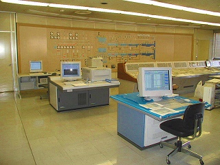 大規模な制御盤が壁に設置され、複数のデスクトップパソコンやプリンターが置かれた、広々とした監視室の様子の写真