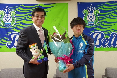 水色と緑を基調としたユニフォーム姿の男性が、スーツ姿の市長から花束とマスコット人形を受け取っている屋内写真