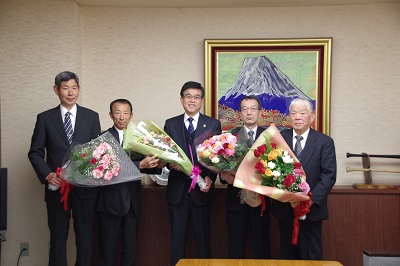 富士山の絵画が飾られた部屋で、市長を中央にスーツ姿の5人の男性が、それぞれ花束を持って並んで立っている贈呈式の写真