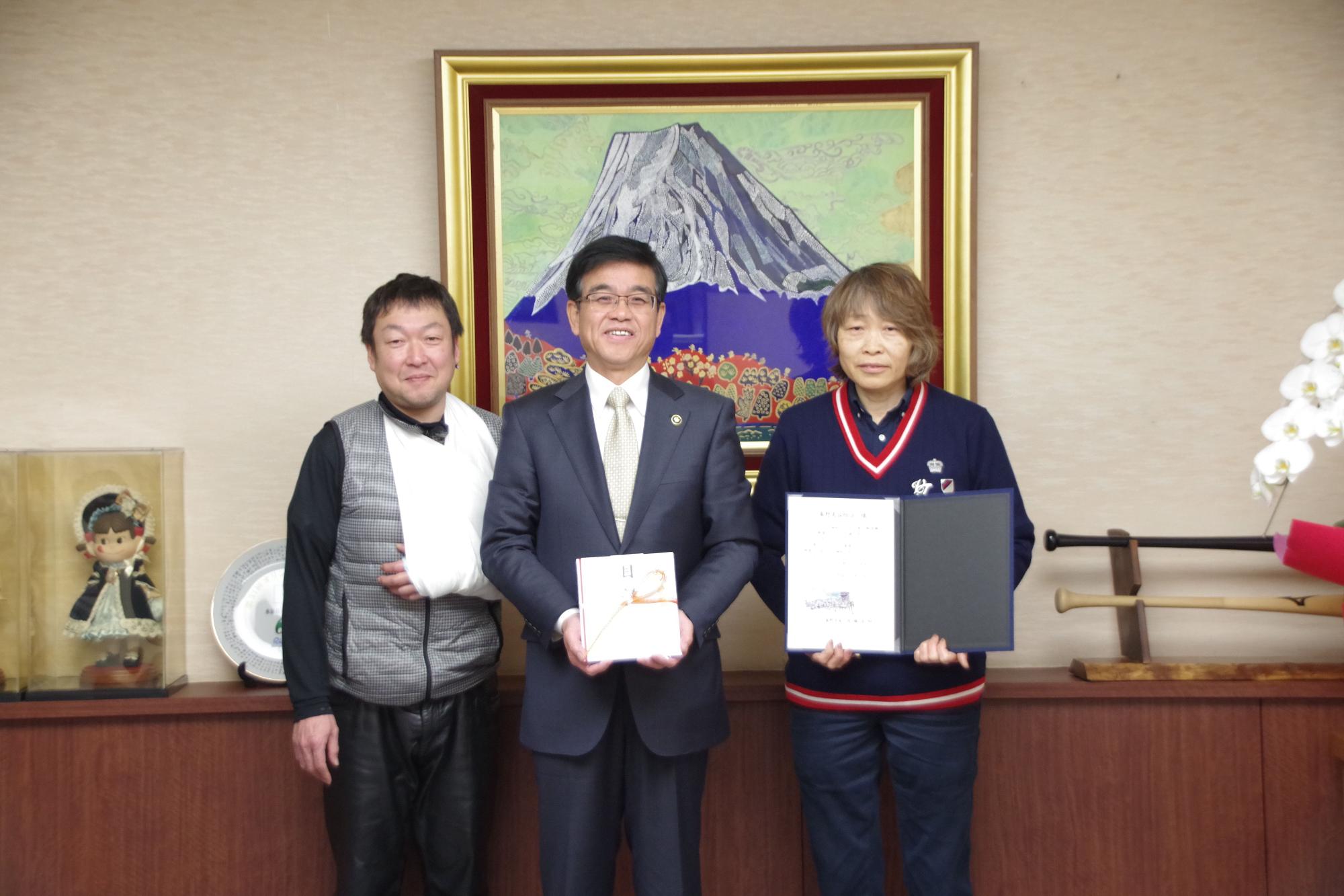 富士山の絵画が飾られた部屋で、スーツ姿の市長が熨斗袋の目録を持ち、両脇を男女の二人に囲まれ、右側の女性は感謝状のような書類を携えている記念写真
