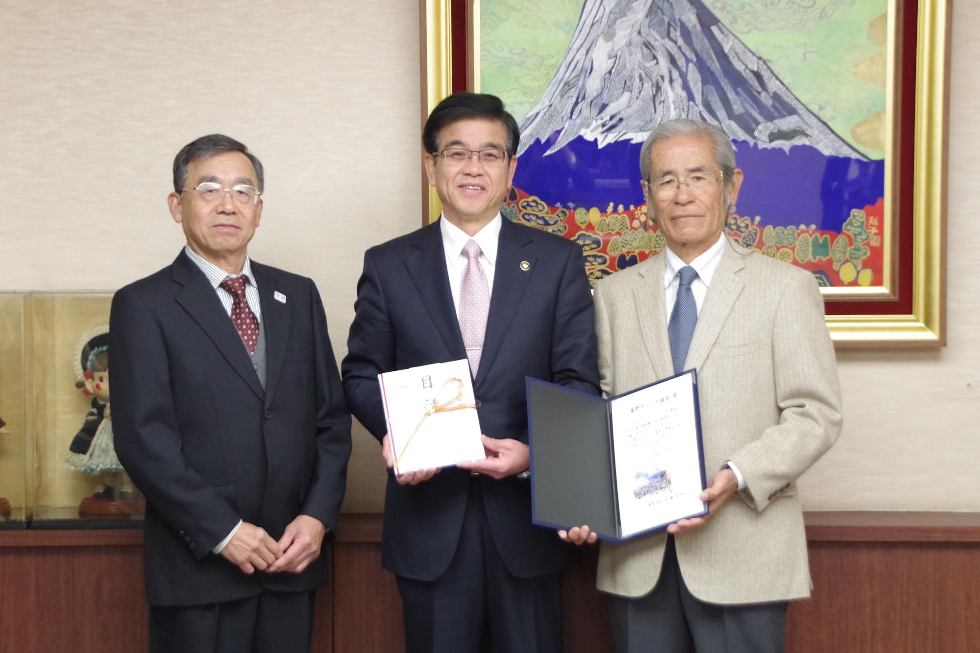 富士山の絵画が飾られた部屋で、市長の両脇に男性が立ち、市長は紅白の熨斗紙の目録、右の男性は感謝状のような書類を持ち、並んで写っている贈呈式の写真