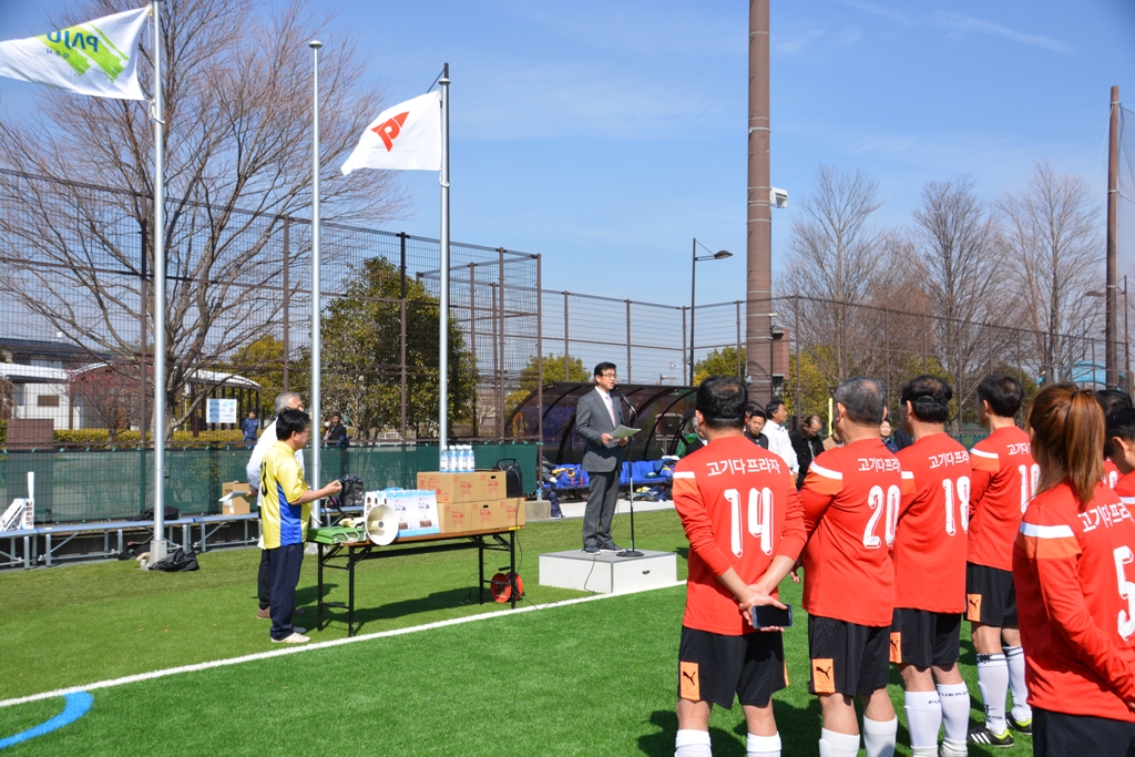 屋外のグランドで、背番号が入った赤いユニフォームを着た選手たちが整列している前で、スーツ姿の市長がマイクを使い挨拶をしている写真