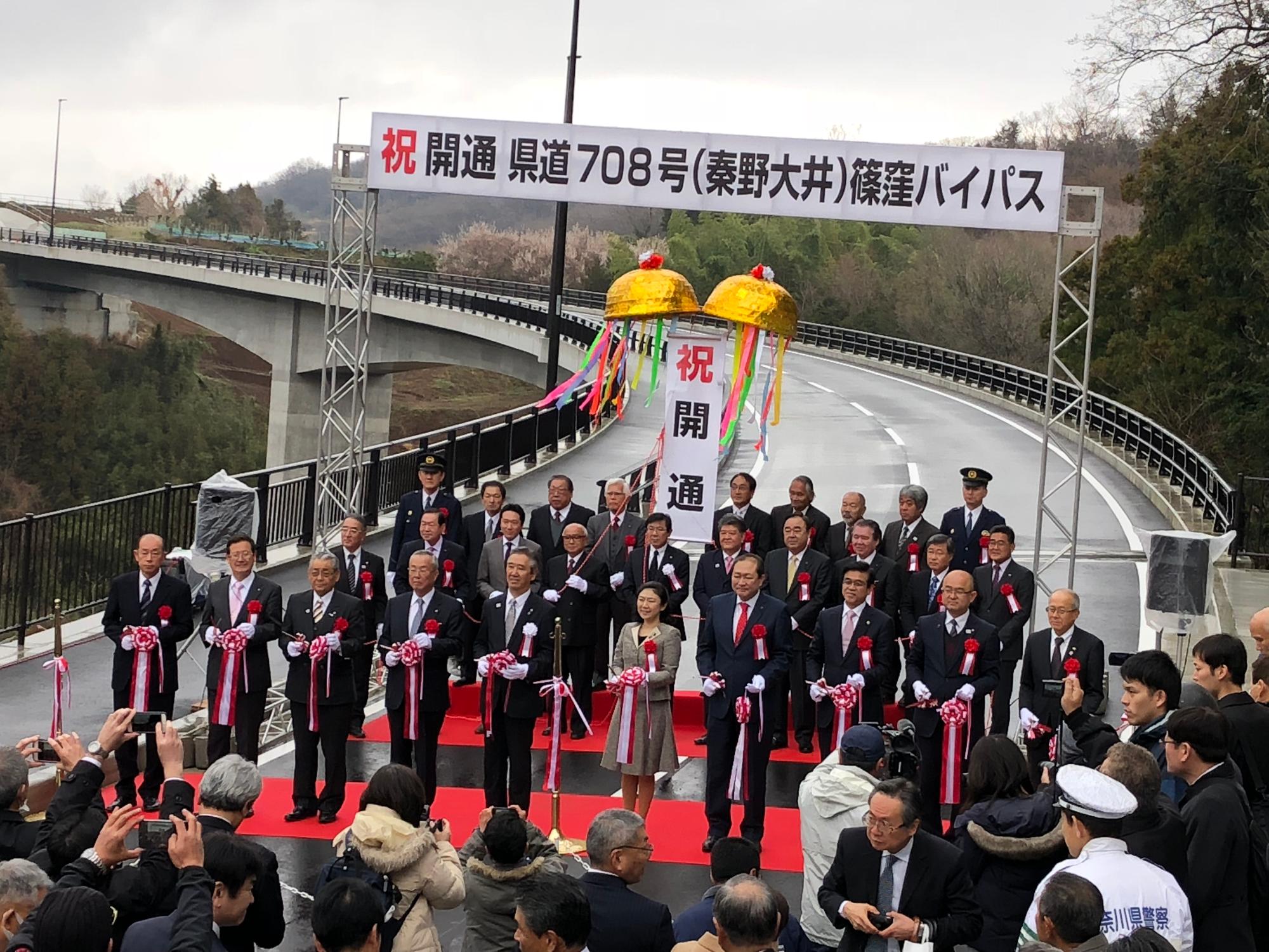 紅白のテープと金色のくす玉が飾られた道路を背景に、赤い絨毯の上で、多くのスーツ姿の男女が並んで立っている開通式の写真