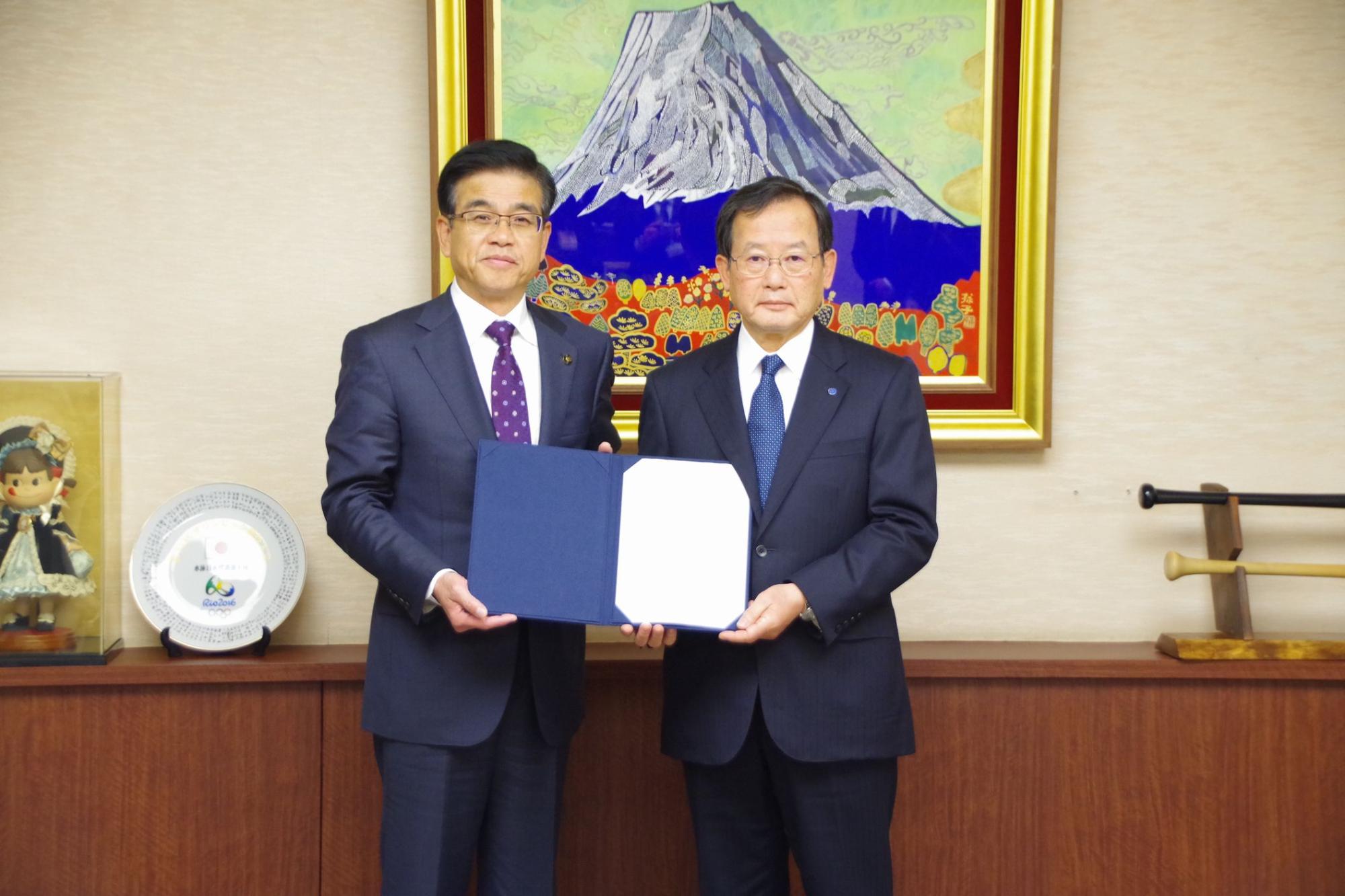 富士山の絵画が飾られた室内で、スーツ姿の市長と男性が、中央で開いた紺色のファイルの書類を持って並んでいる写真