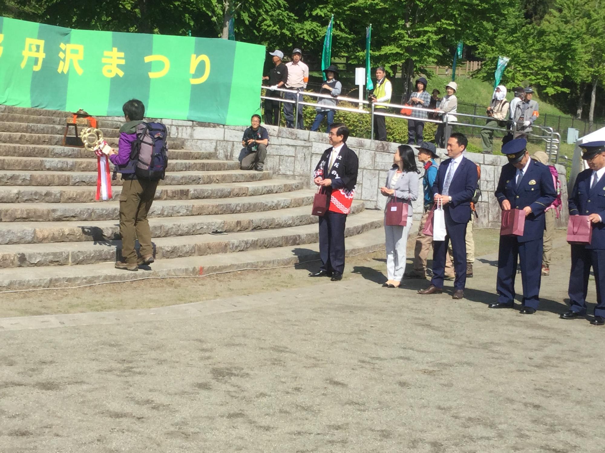 「秦野丹沢まつり」の横断幕が階段横に掲げられており、関係者が見守る中、リュックを背負った登山客が階段を登ろうとしている写真