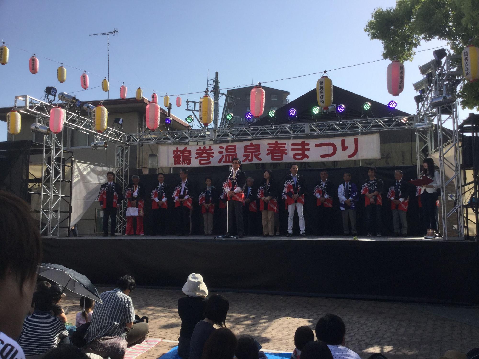 「鶴巻温泉春まつり」の横断幕が掲げられた祭りの特設ステージ上に、法被姿の複数の人々が並んでおり、中央に立ちマイクスタンドで話をしている高橋市長の写真
