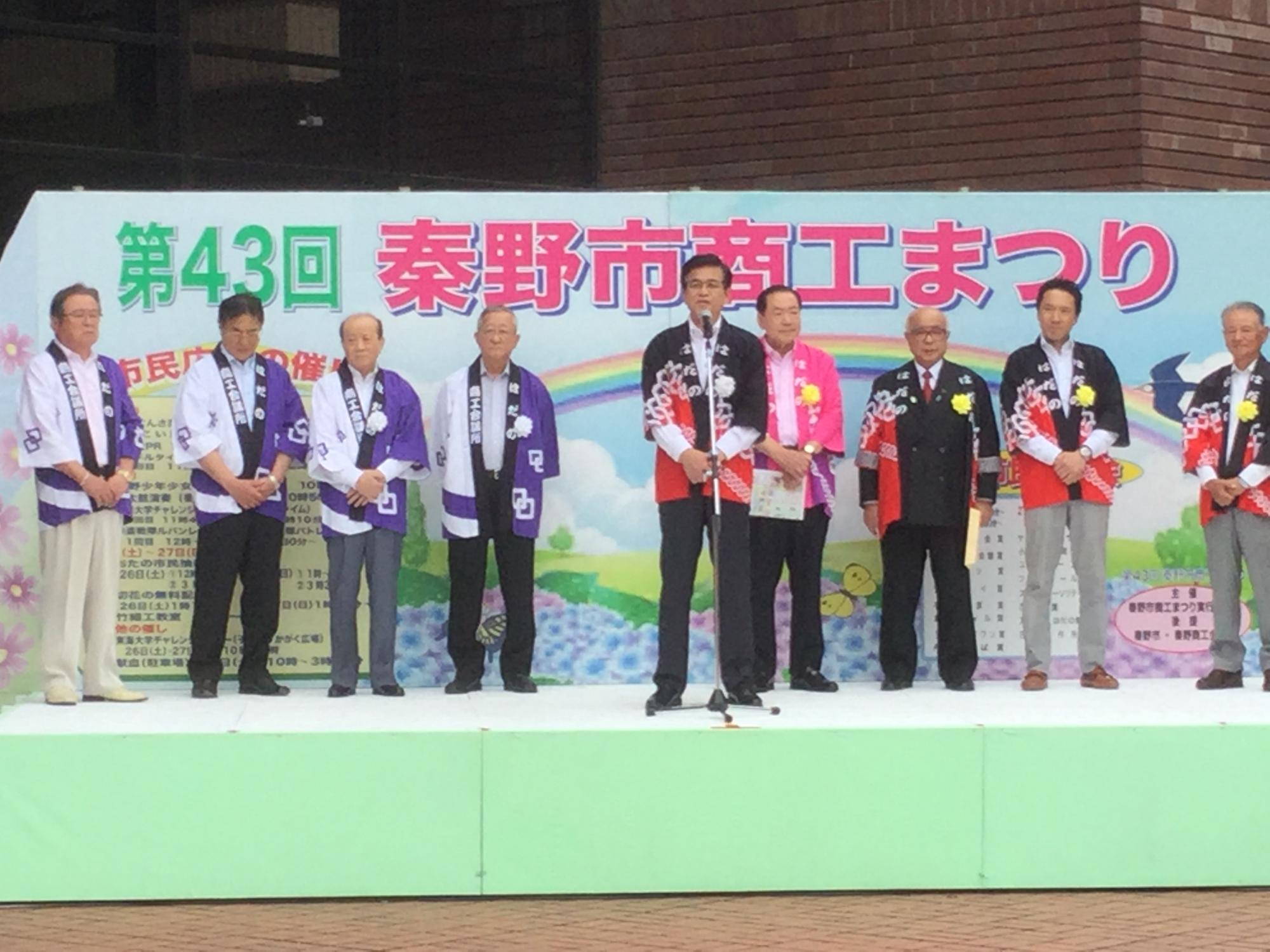 屋外ステージに立ち、法被を着た関係者と、マイクを前に高橋市長がスピーチをしている写真