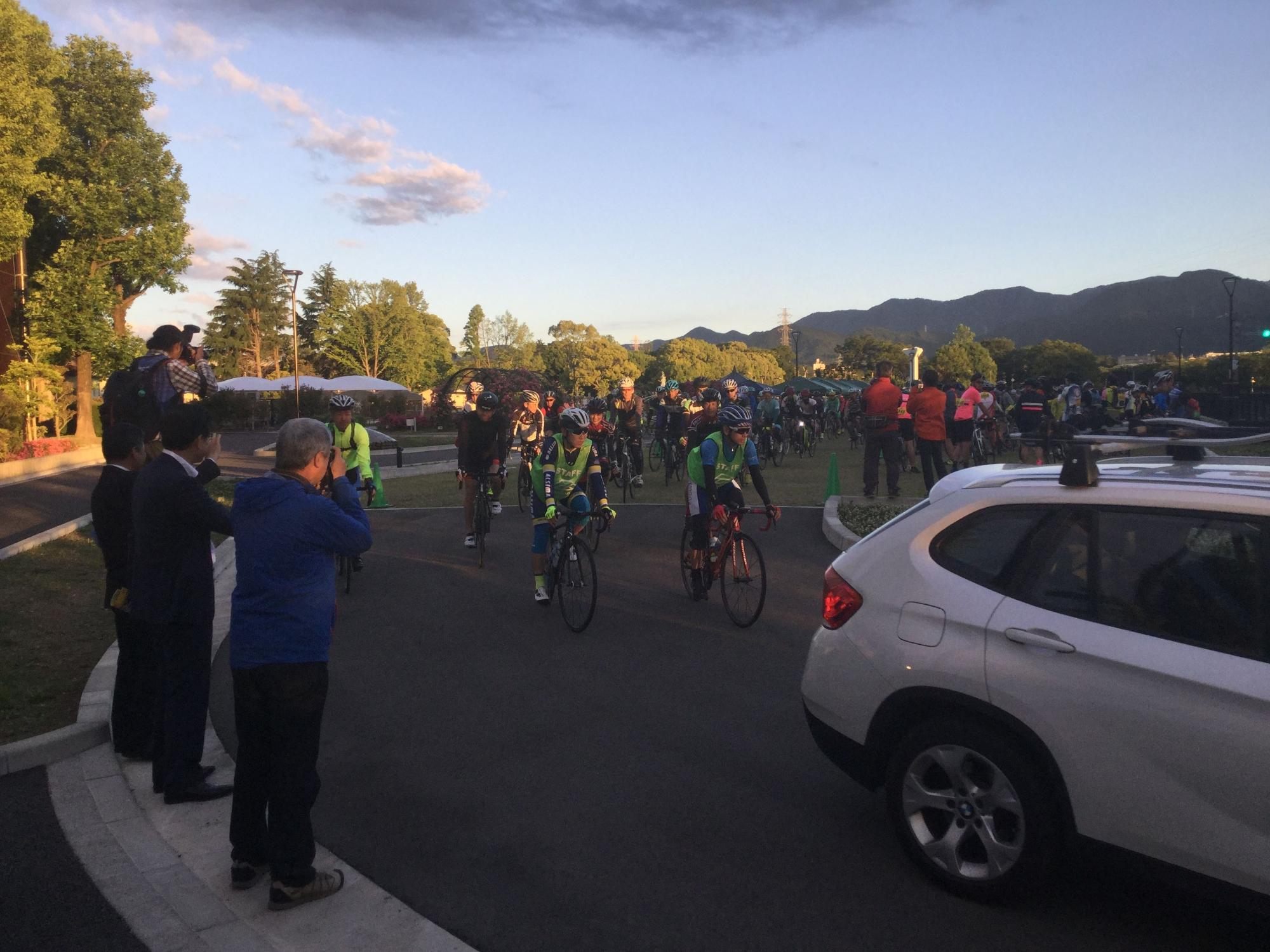 早朝に出発するロードバイクに乗った参加者と、それを眺める関係者と観客の写真