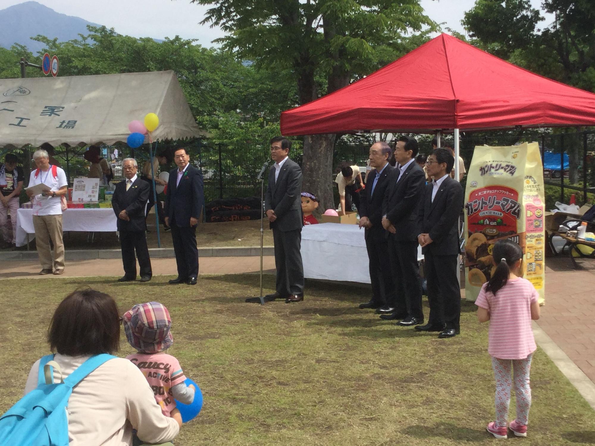 晴天の下、親子連れや子どもが集まる公園に赤と白のテントが設営され、高橋市長がスーツ姿の関係者と並び、マイクスタンドの前で話している写真