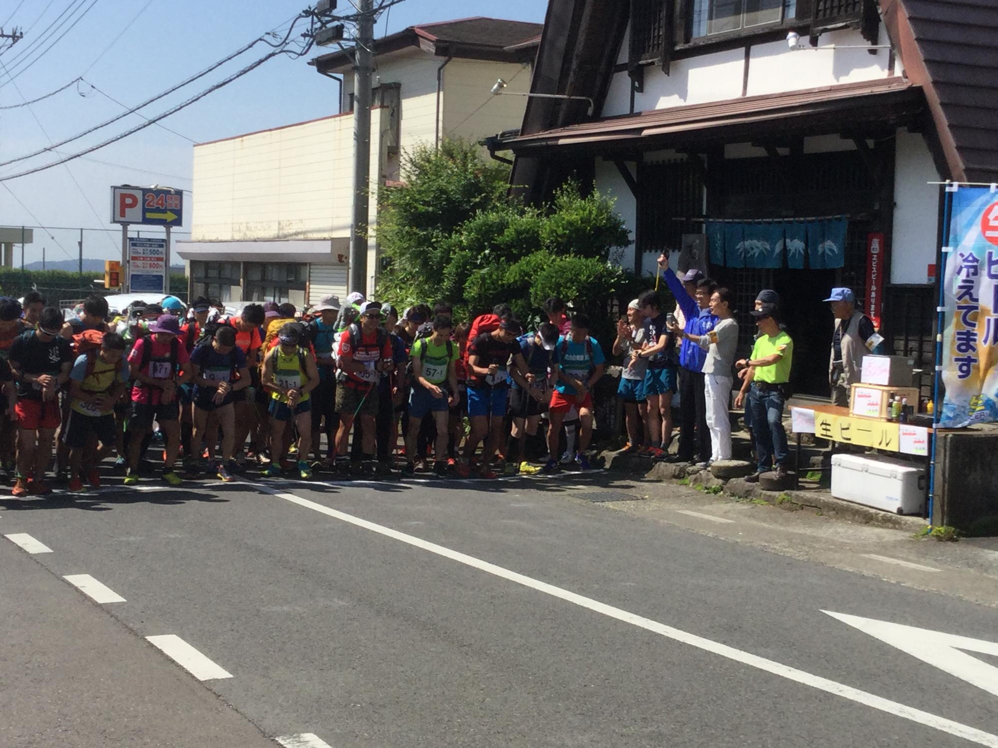 沿道で人々が見守る中、歩荷を背負った大勢のランナーたちが道路のスタート地点に集まり、スターターピストルを空に向けて構えた高橋市長のスタートの合図を待っている様子の写真