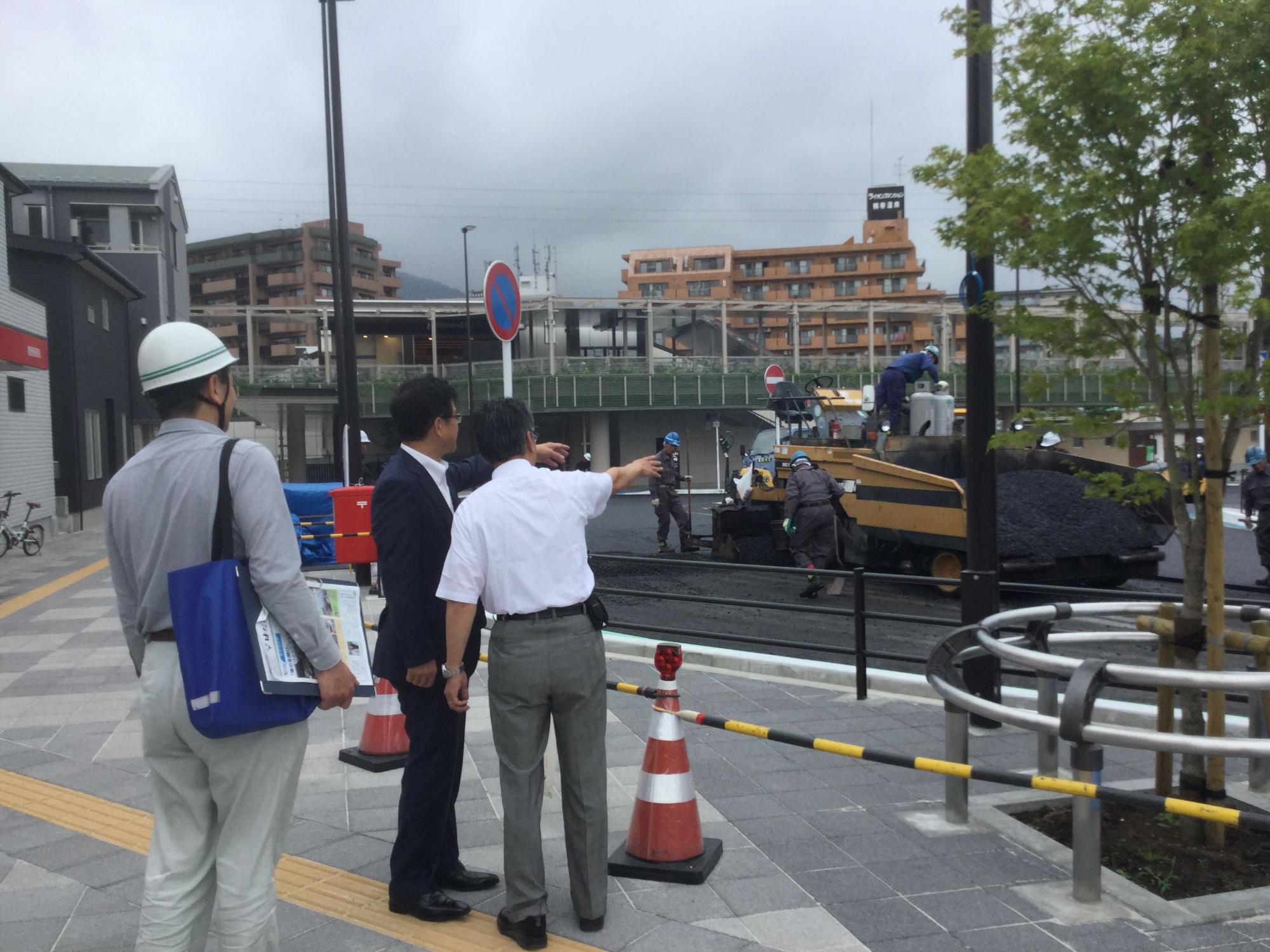 鶴巻温泉駅南口広場の通りで工事車両による舗装工事が行われいる様子を、少し離れた歩道から高橋市長と関係者男性が指をさしながら確認しており、その後ろにヘルメットをかぶった男性が立っている写真