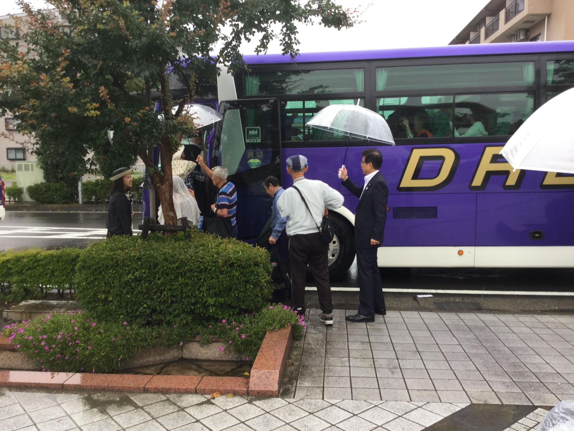 道路の路肩に紫色の車体の大型バスが停車しており、歩道の植栽の前からバスに乗り込んでいく参加者たちを、手に持った傘を差しだしながら高橋市長が見守っている様子の写真