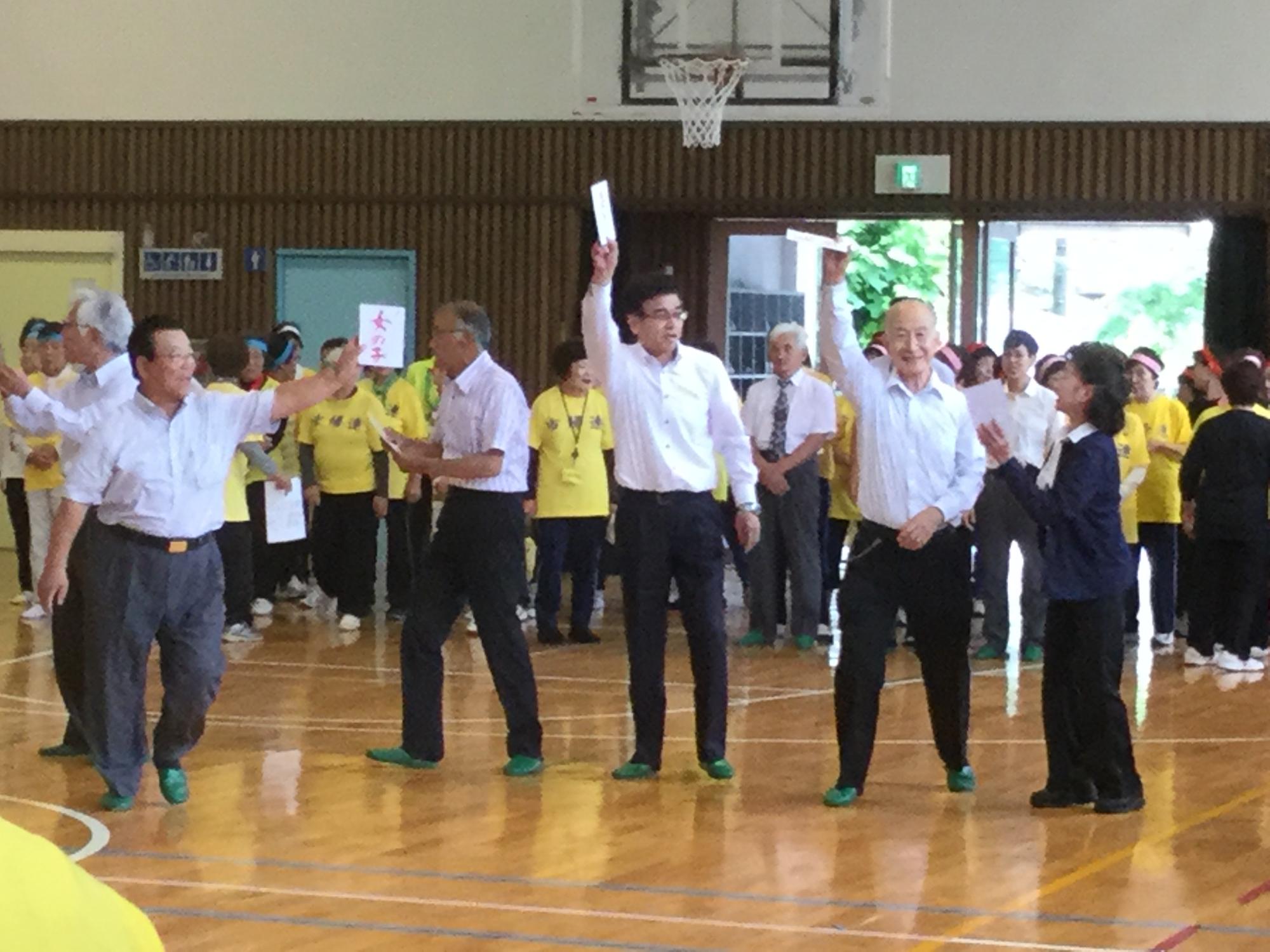 体育館で、年配の男女が紙を掲げて相手探しのゲームを行っており、楽しそうにレクリエーションに参加する高橋市長と参加者の写真