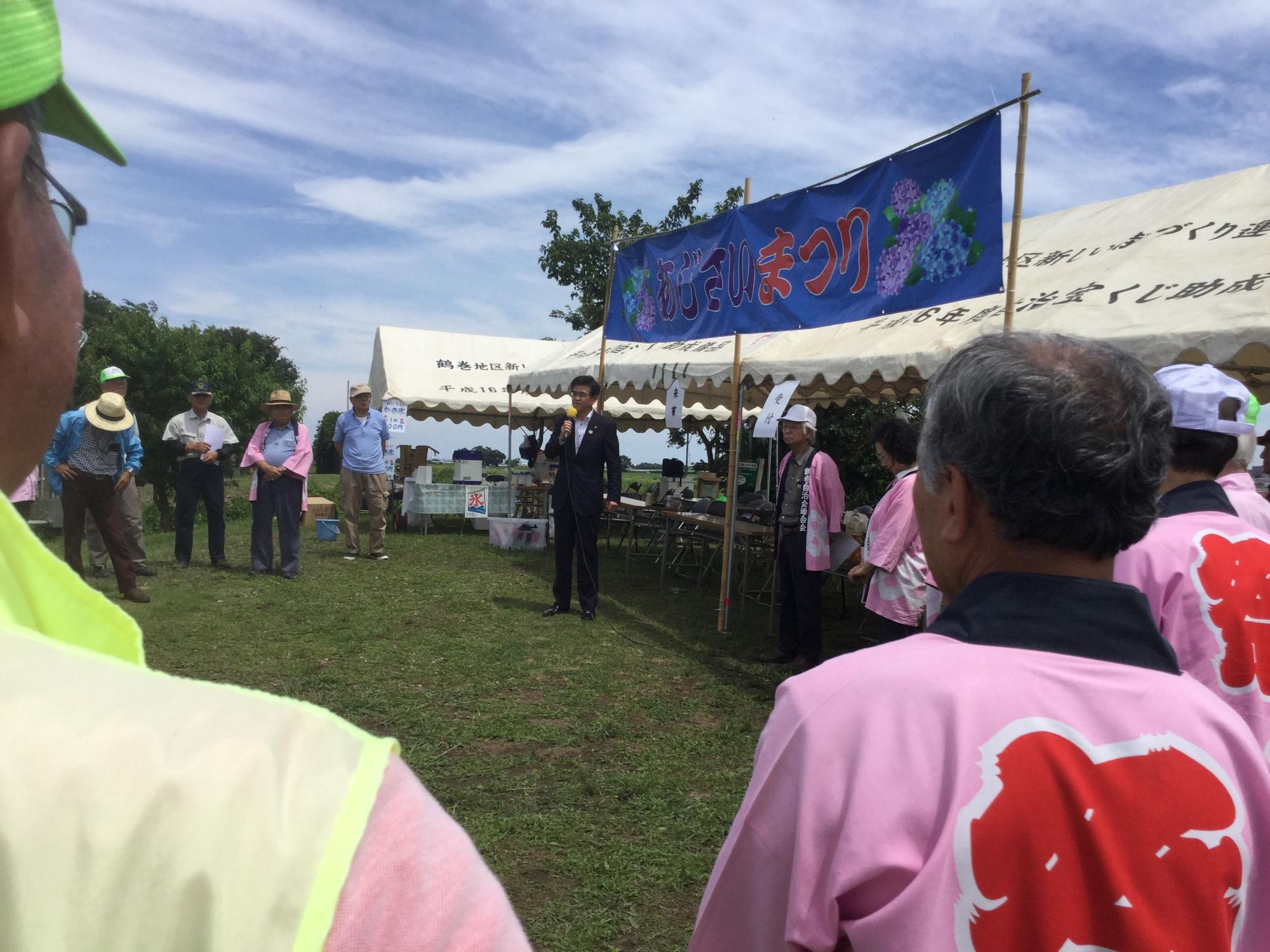 芝生の広場に、複数のテントが並び、「あじさいまつり」の横断幕の下に立った高橋市長が、輪になって立ったピンク色の法被を着た関係者などに向かって話をしている写真