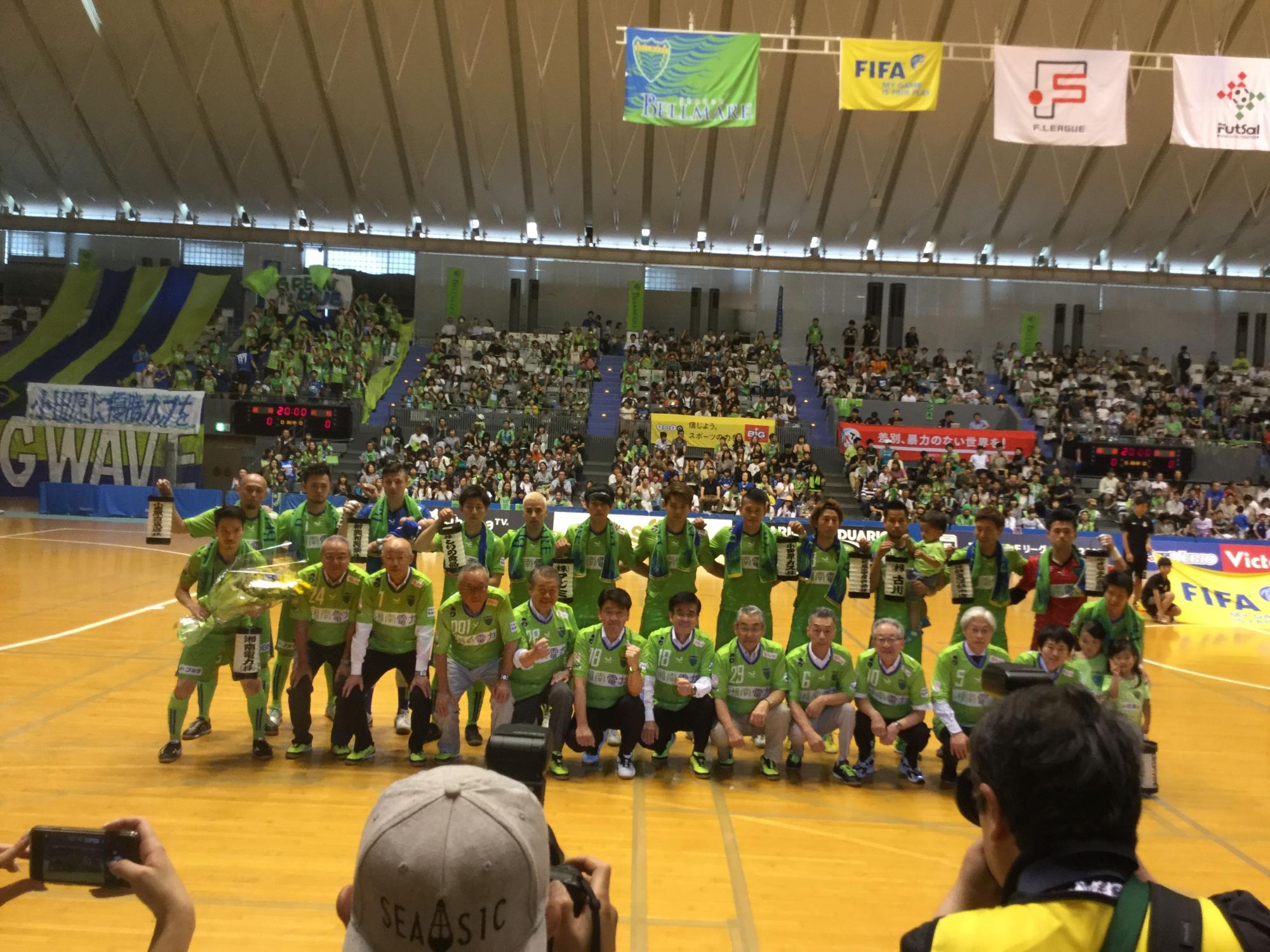 大勢の観客が入っているアリーナのコート上で緑色のユニフォームを着たチームメンバーと高橋市長が前後2列に並んで記念撮影をしている写真