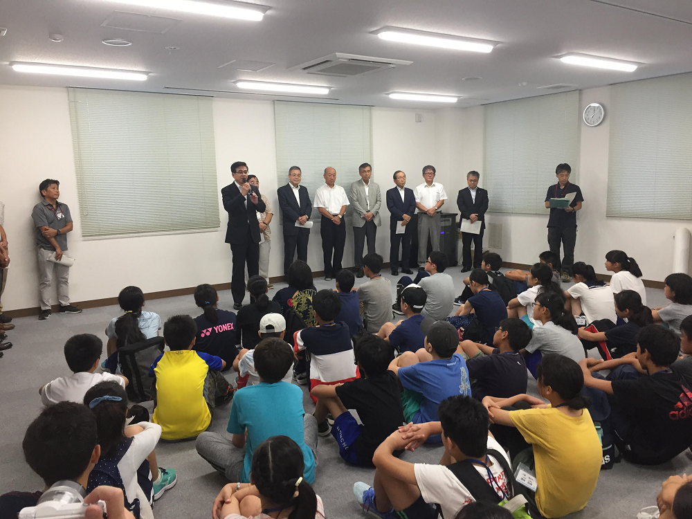 窓がブラインドで覆われた会議室のような広い部屋で、たくさんの子供たちが床に座り、前方の壁側に複数の関係者の男性たちが横並びに立つ中、中央に立った高橋市長がマイクを持って話をしている写真