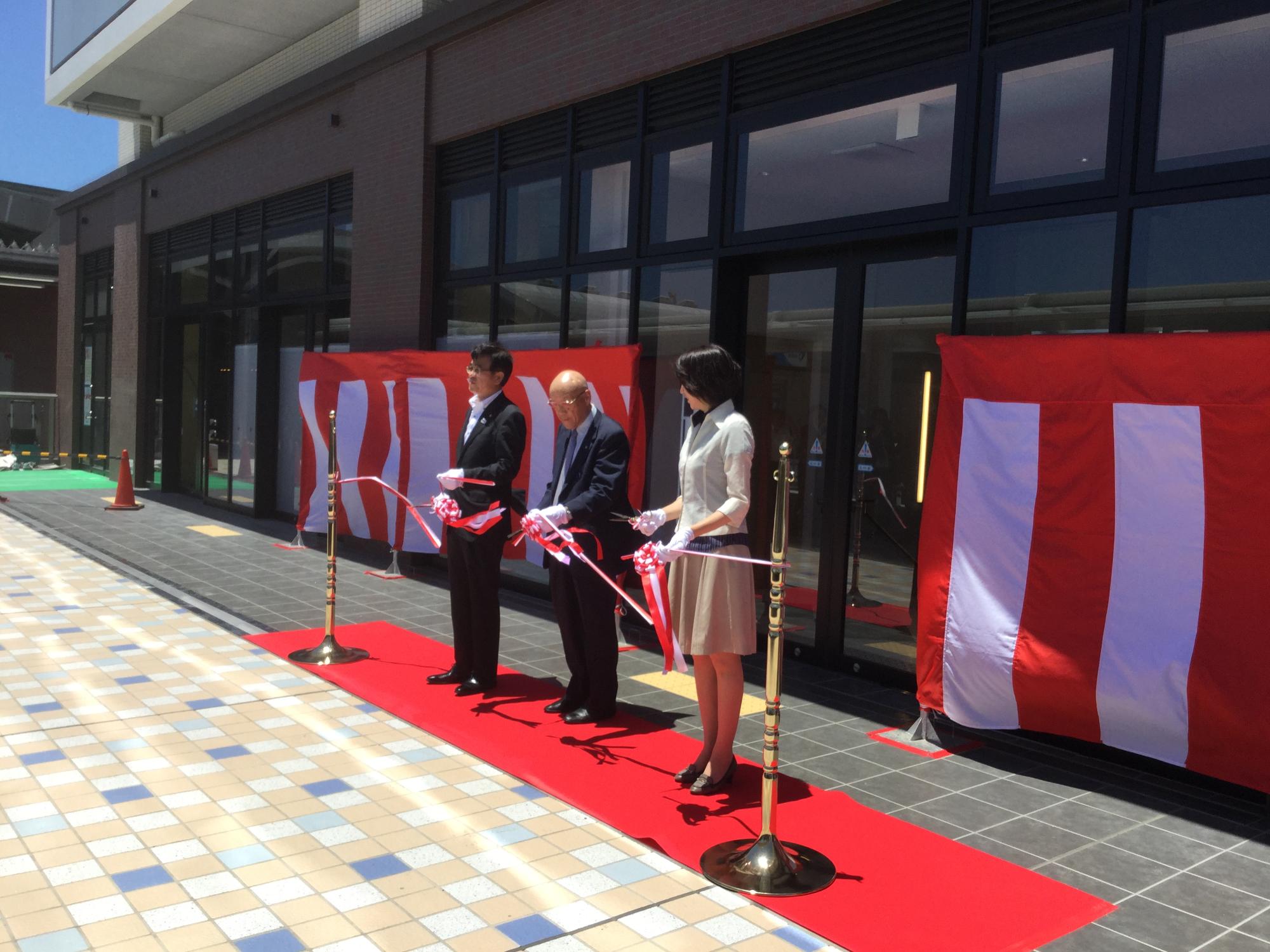 紅白幕が張られた新しい建物の入り口前で、赤いカーペットの上にスーツ姿の市長と関係者の男女が並んで立ちテープカットのセレモニーが行われている様子の写真