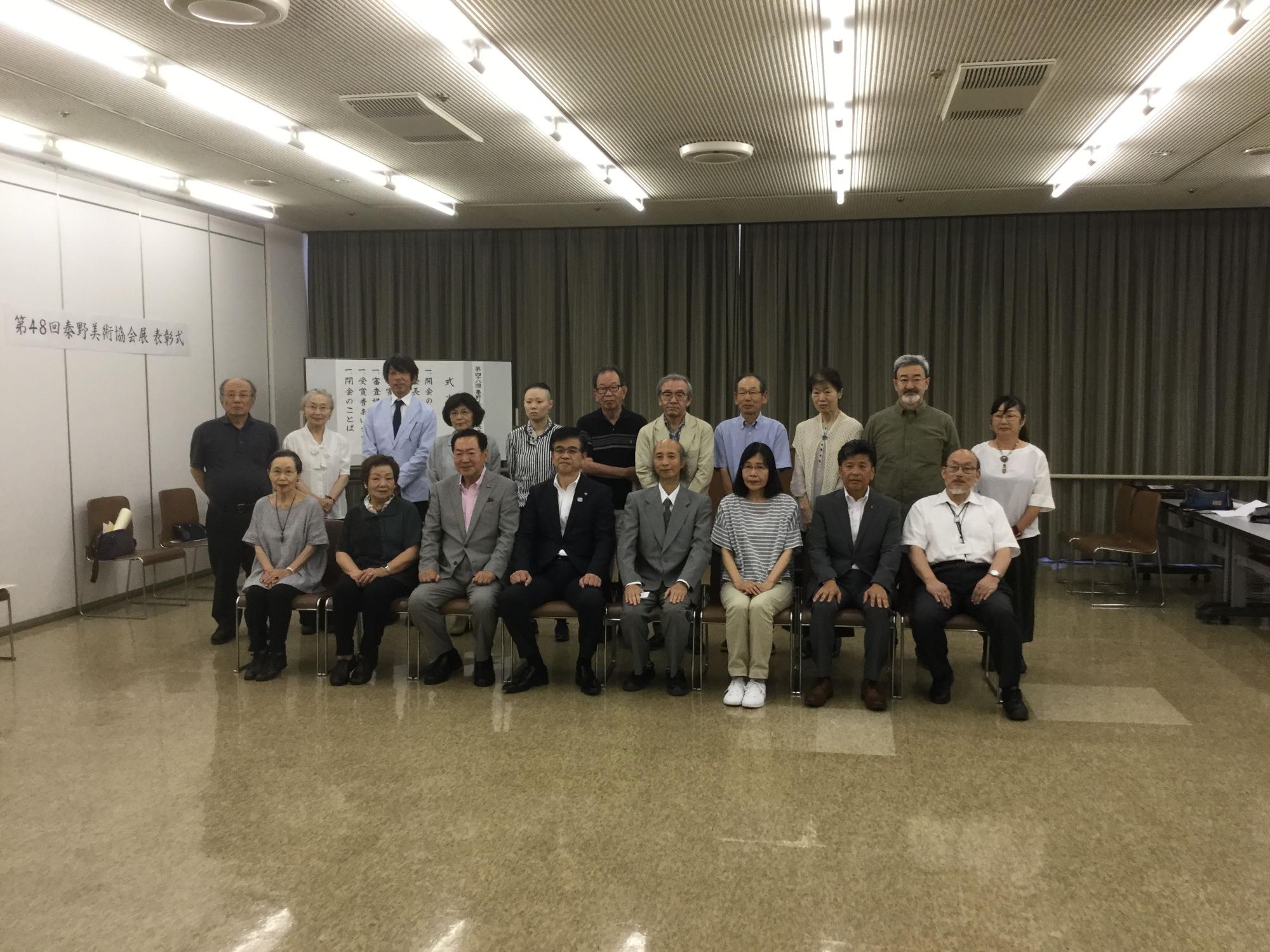 広い室内で、カーテンを閉めた大きな窓を背後に、市長と男女合わせて十数名ほどのグループが、前列は市長を中央にして椅子に座り、後列は立って写した集合写真