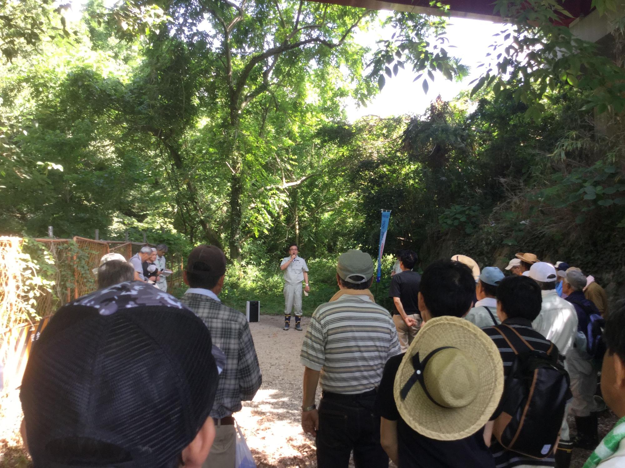 木々が生い茂る屋外の広場に集まった、帽子などをかぶった大勢の参加者が、中央でマイクを持って立っている作業着姿の市長の話を聞いている様子の写真