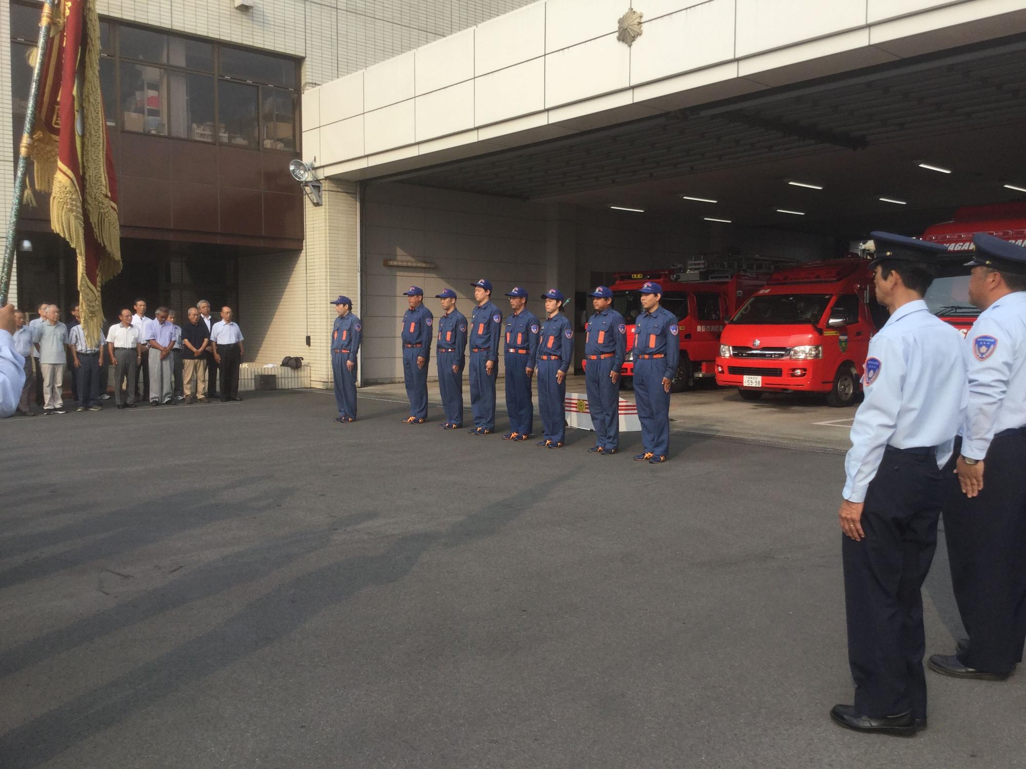消防者などの車両が停められた消防署の車庫の前に制服姿の8人の隊員が横一列に整列し、その左右に職員や関係者たちが整列している壮行会の様子の写真