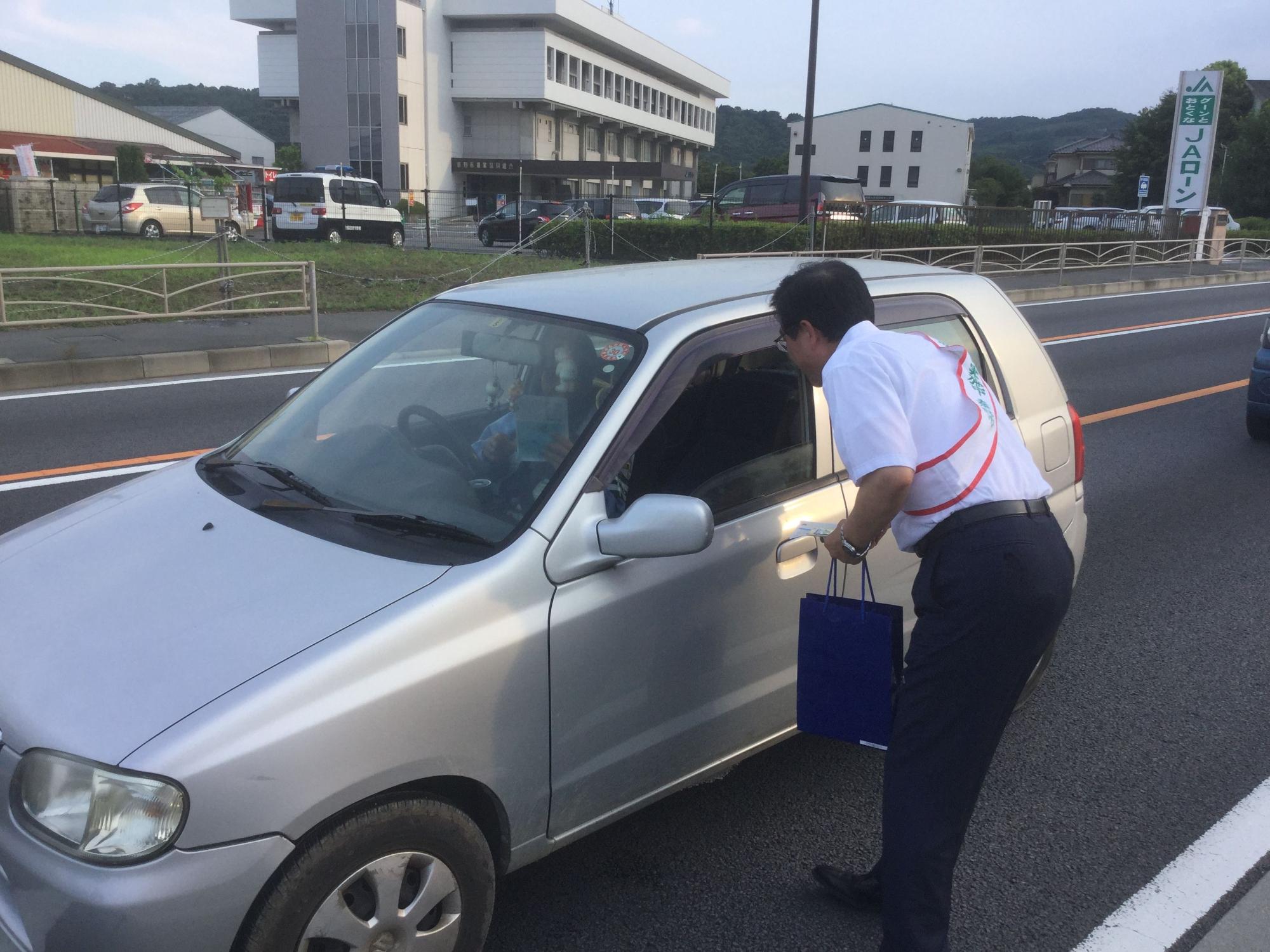 複数の建物や駐車場が奥に見える道路で、白いワイシャツを着て赤い縁取りの白の襷をかけ、青い紙袋を持った市長が、シルバーの自動車の窓越しに運転手と話をしている写真