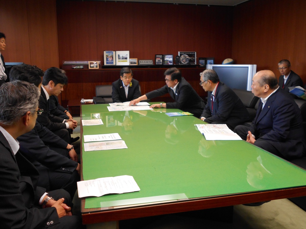 緑色の大きなテーブルを囲んで、複数のスーツ姿の男性たちが資料を広げて座り、中央の席に座った男性の前に広げた資料を指さしながら高橋市長が説明をしている様子の写真