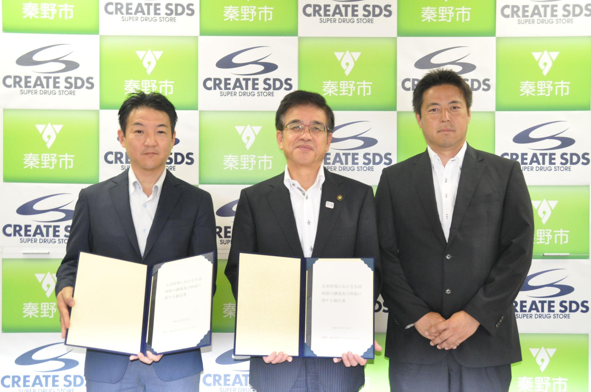 緑と白のチェッカー模様のバックパネルの前で、スーツ姿の市長と男性2人が、協定書を持って並んで立っている記念写真