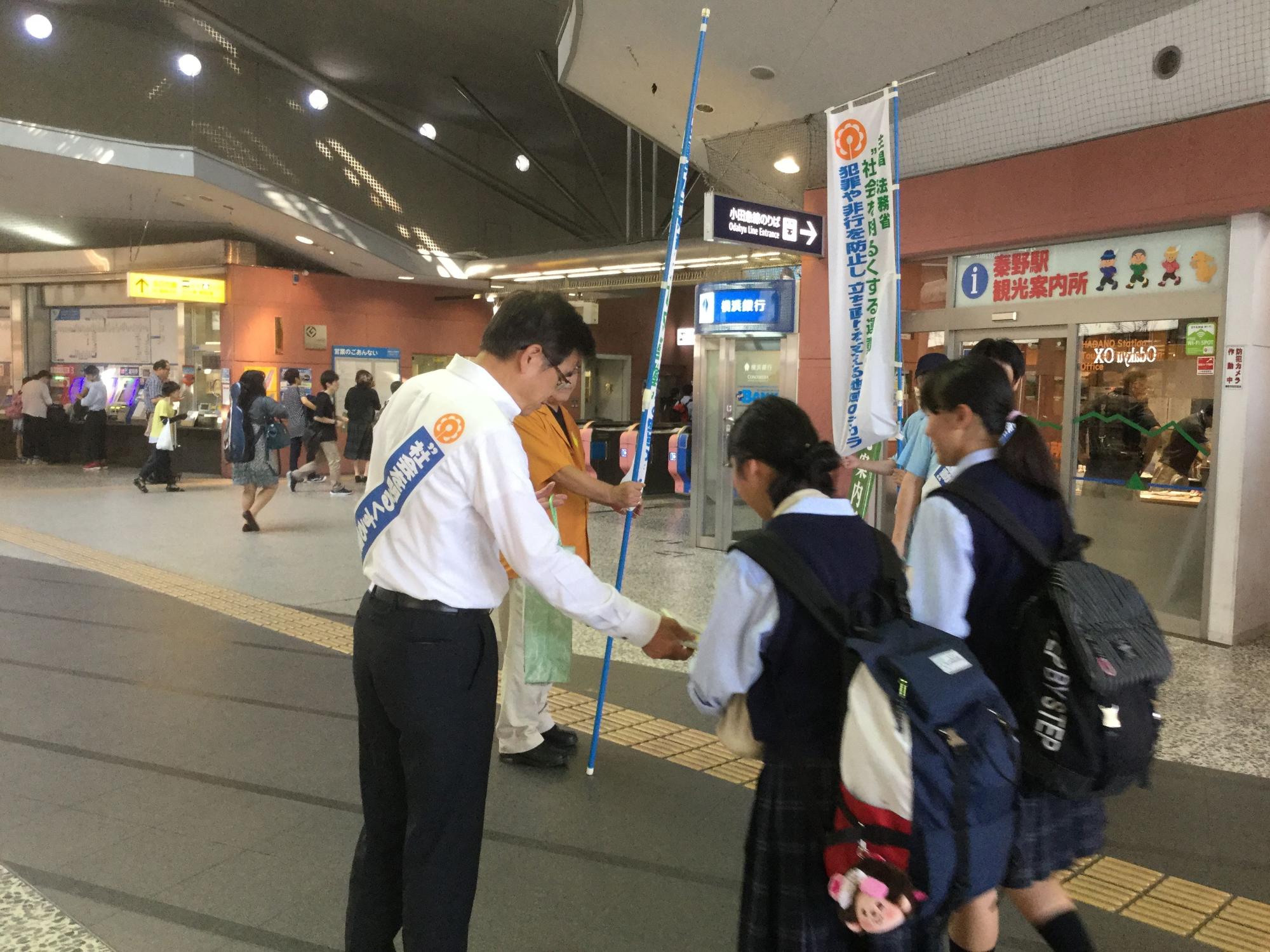 駅の構内で、啓発運動の襷をかけた市長が、制服姿の2人の女子高校生に資料を手渡している様子の写真