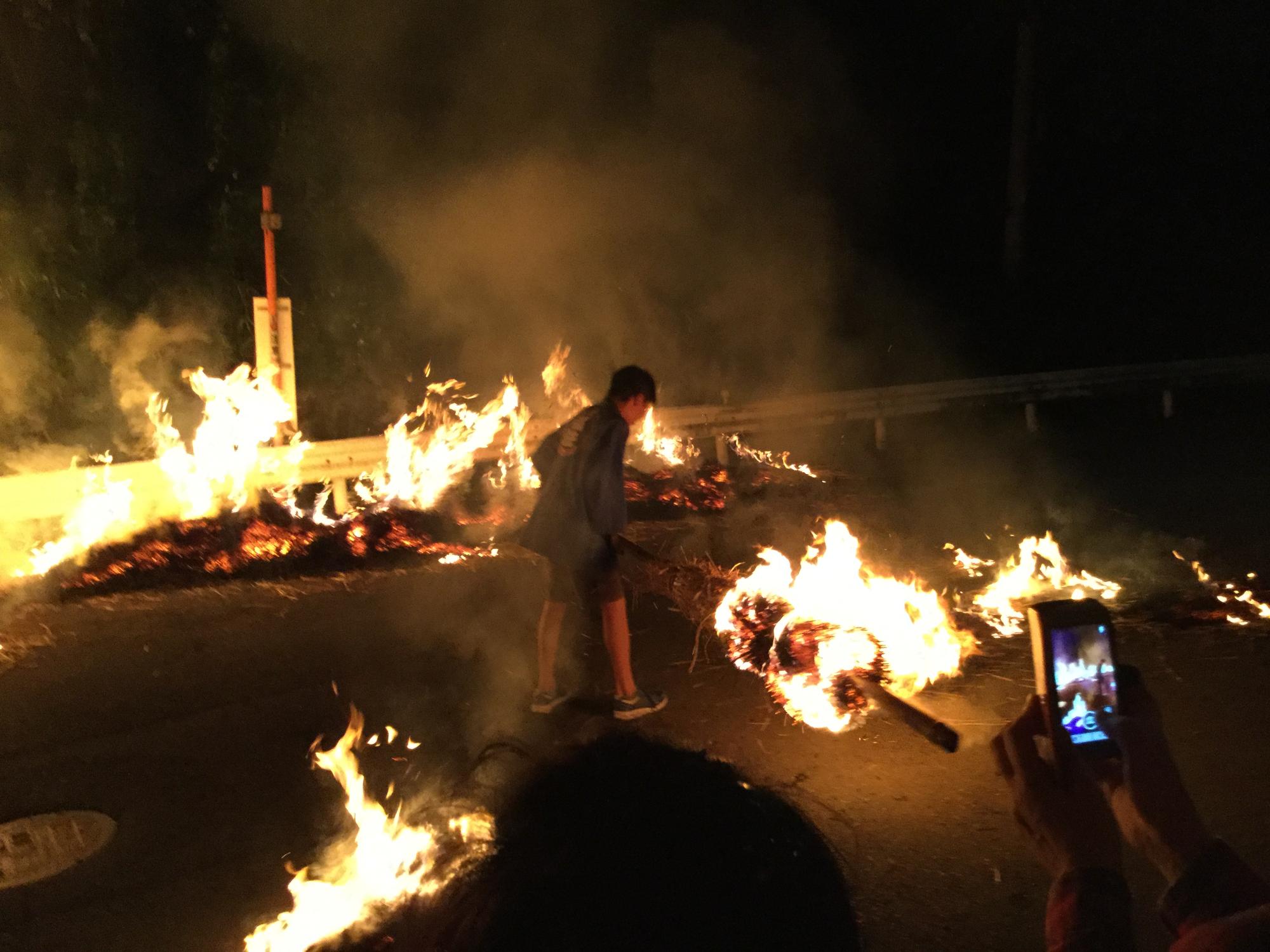 夜の路上で、一人の男性が燃え盛る火に長い松明を振り回しており、周囲には炎と煙が立ち込めている「瓜生野百八松明」の様子の写真