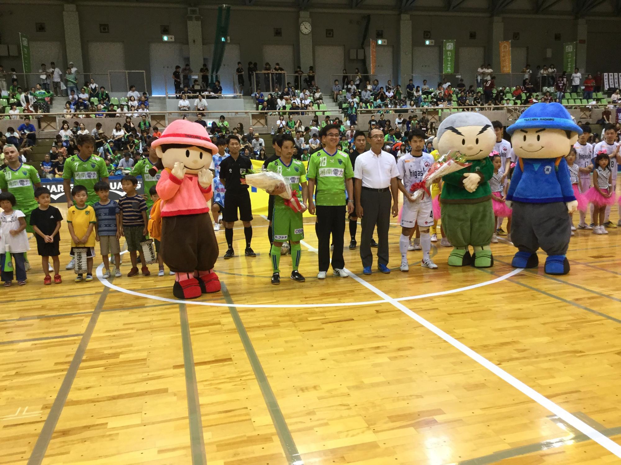 客席に観客が座っている総合体育館で、ユニフォーム姿の高橋市長や子供達、チームメンバーが並び、丹沢はだの三兄弟と一緒に記念撮影している写真