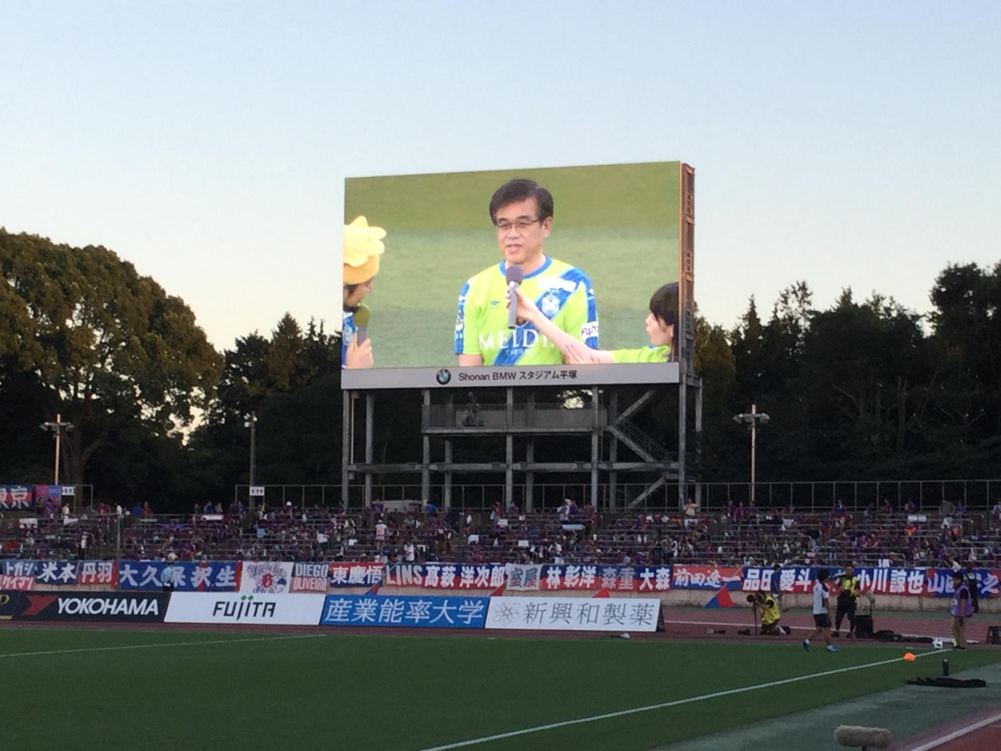 夕暮れの中、多くの観客が座っているスタジアムの大型ビジョンに、緑色のユニフォームを着てマイクで話をする高橋市長が映し出されている写真