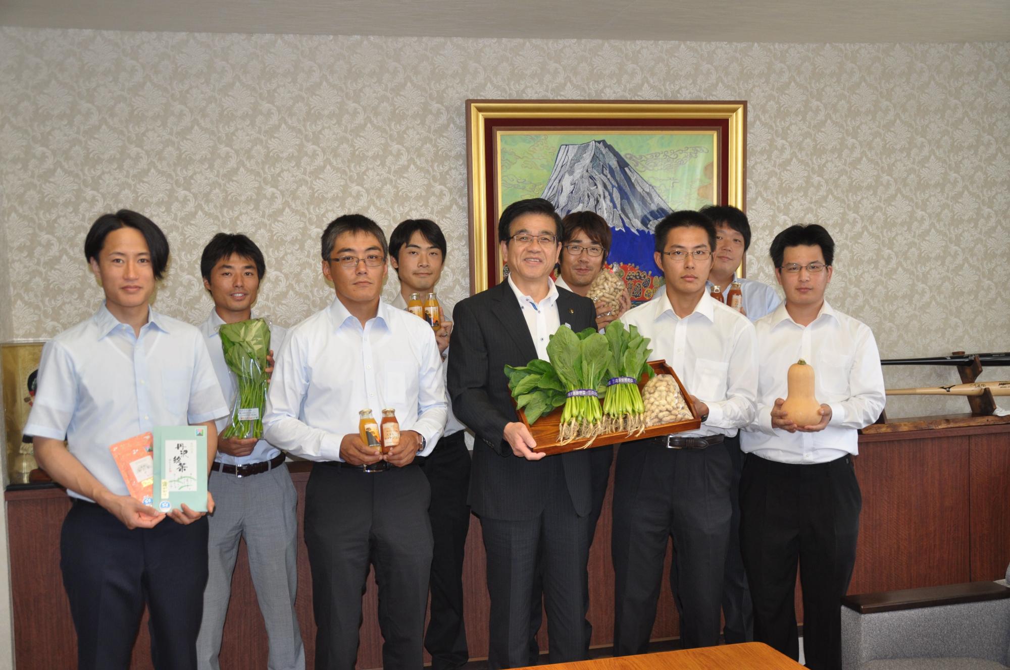 応接室のような部屋で、白いシャツを着た複数の男性達と高橋市長が地元の野菜や加工品を手に持ち、記念撮影を行っている写真