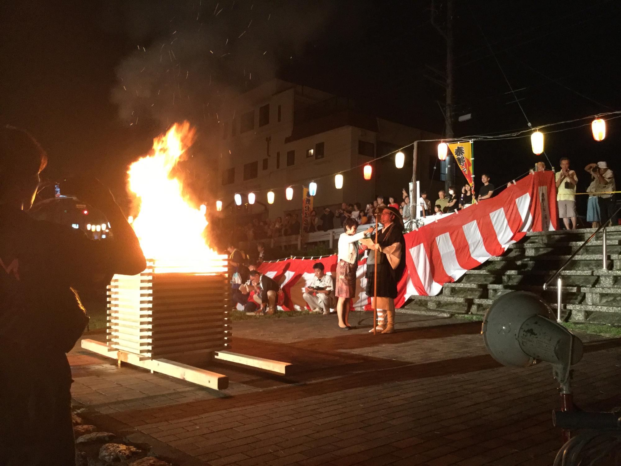 激しく燃え上がる大きな焚き火の前で、話をする高橋市長と、沿道に集まる観覧客の写真