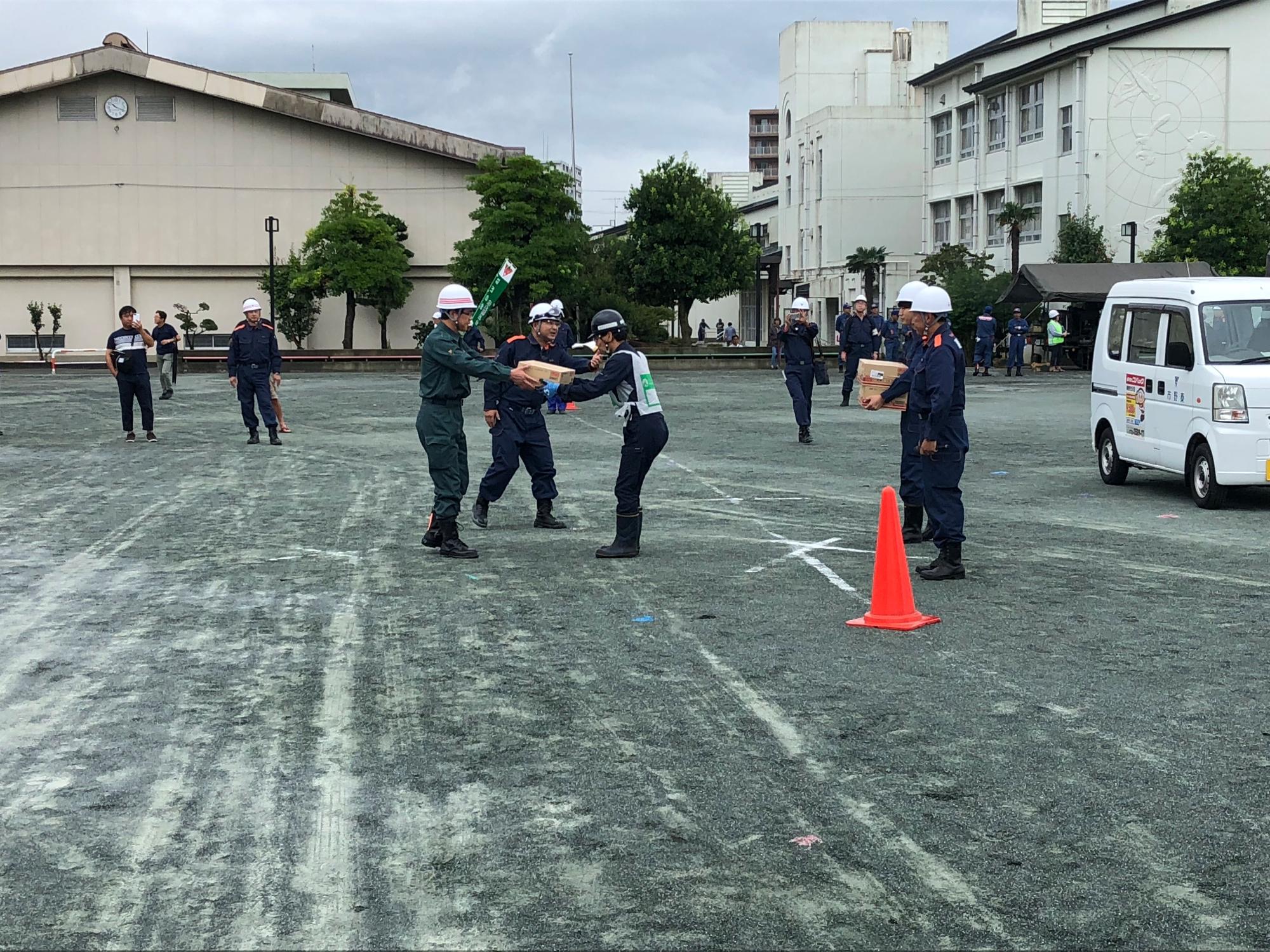 小学校の運動場で、ヘルメットをかぶった参加者が箱を運んで防災訓練を行っている写真