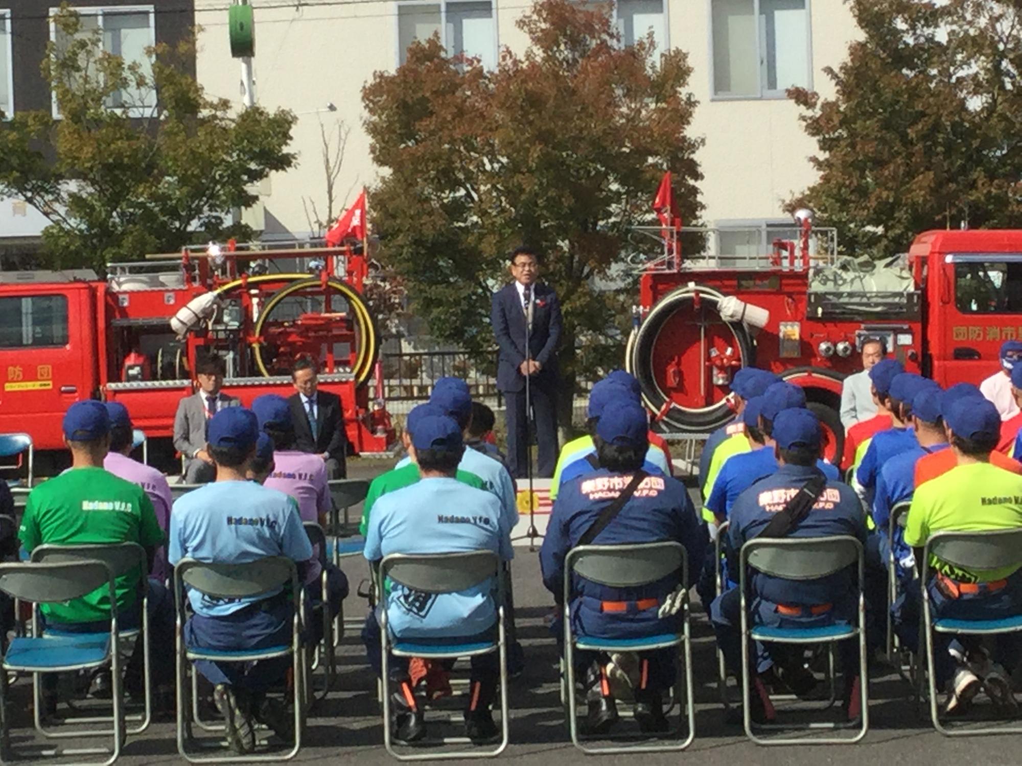 晴天の下、消防団員が椅子に座り、消防車の前で話をする高橋市長の話を傾聴している写真