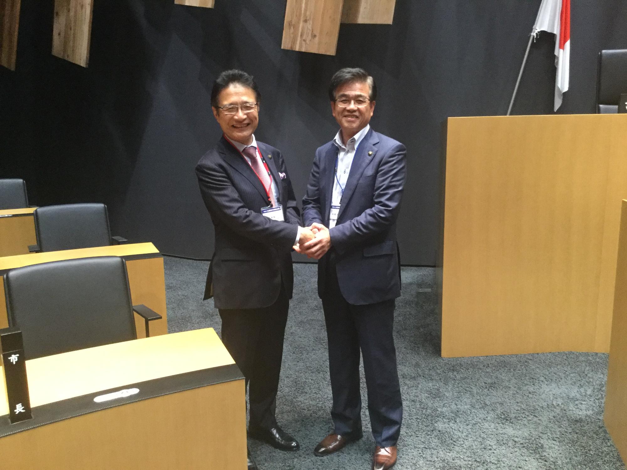 木目調の机と椅子が並ぶ会場で、高橋市長と前葉市長が笑顔でがっちりと握手をしている写真