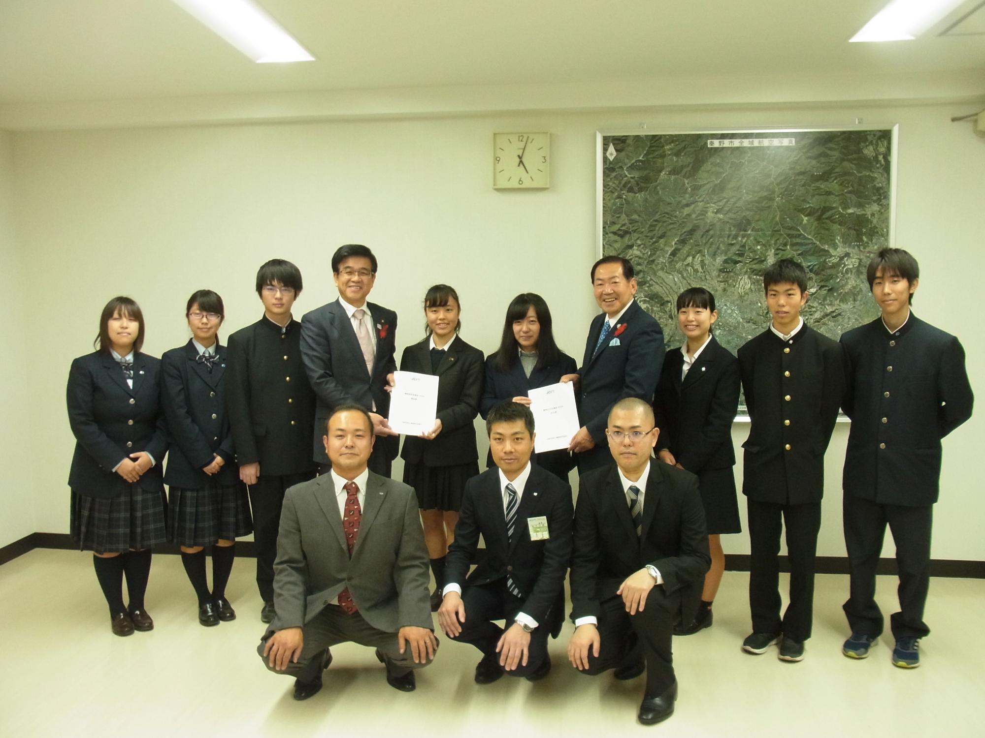 ブレザーや学ランを着た学生とスーツ姿の市長と関係者の大人たちが、賞状のような白い紙を中央に持って集合している記念写真