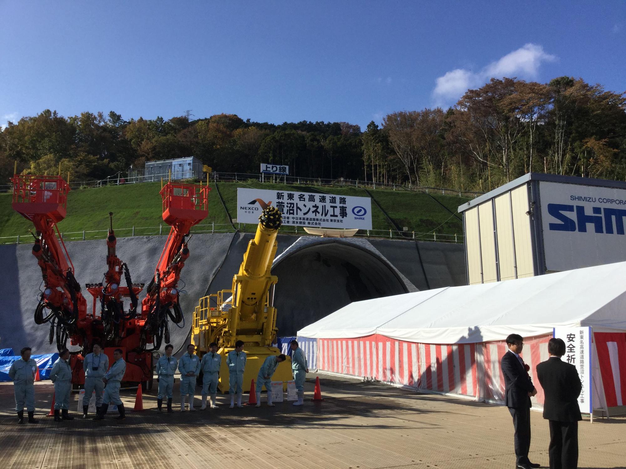 新東名高速道路のトンネル工事現場の前に、オレンジ色の巨大な重機と黄色のクレーンが並び、青い作業服の作業員とスーツ姿の男性たちが集まっている様子を捉えた写真