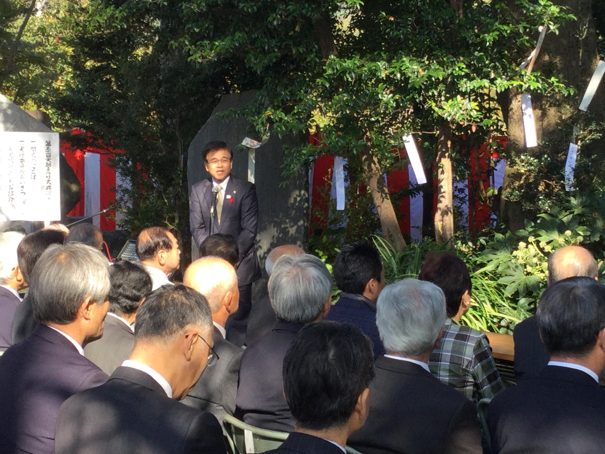 野外の木々が生い茂る場所で、黒いスーツにネクタイ姿の市長が、多数の参加者を前に立った状態で演説を行っており、背景には紅白幕と白い短冊のようなものが木に結ばれている様子が写っている写真