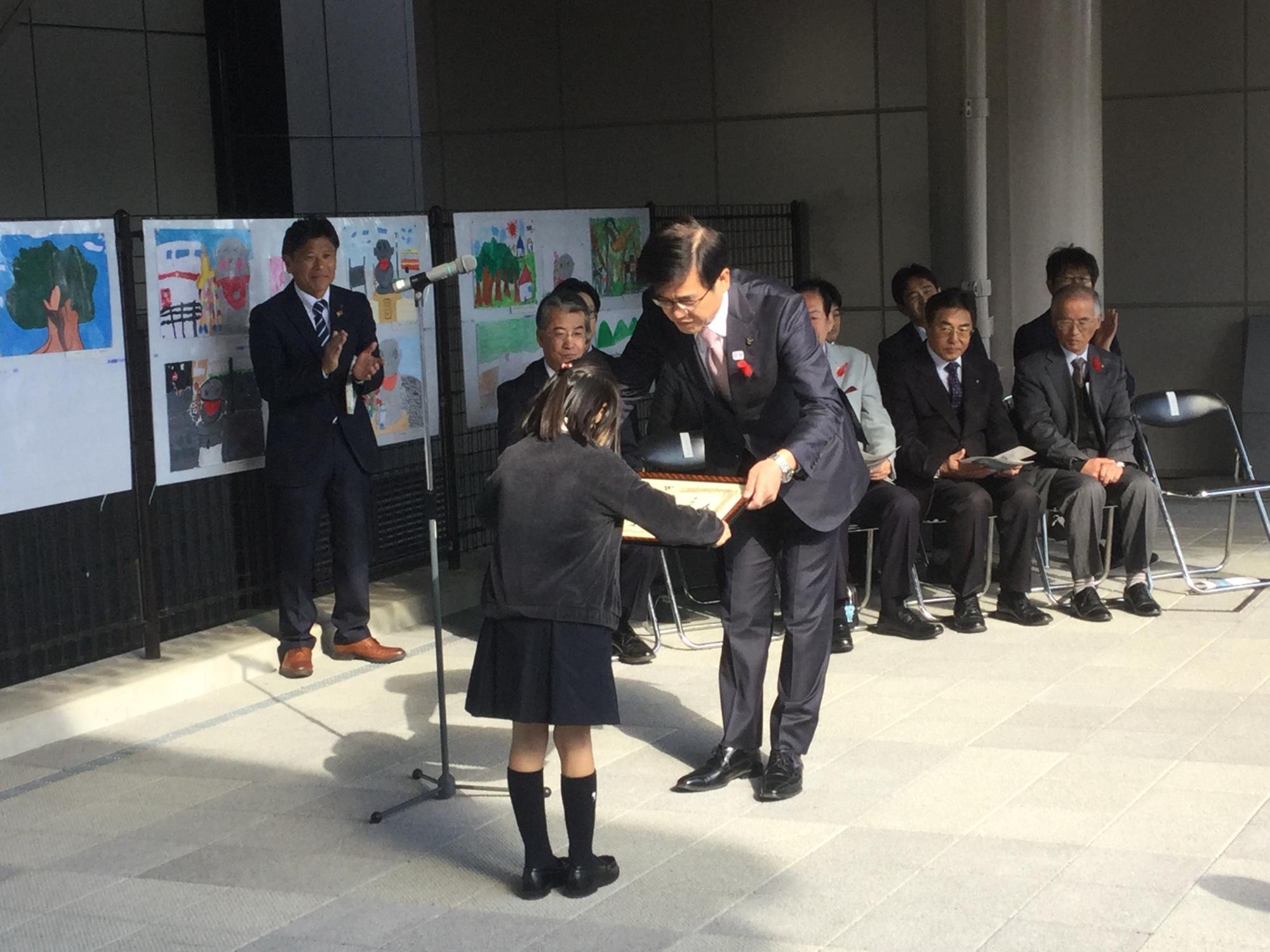 紺色のスーツを着た男性が黒いスカートとジャケットを着用した女の子に、賞状を両手で授与している表彰式の写真