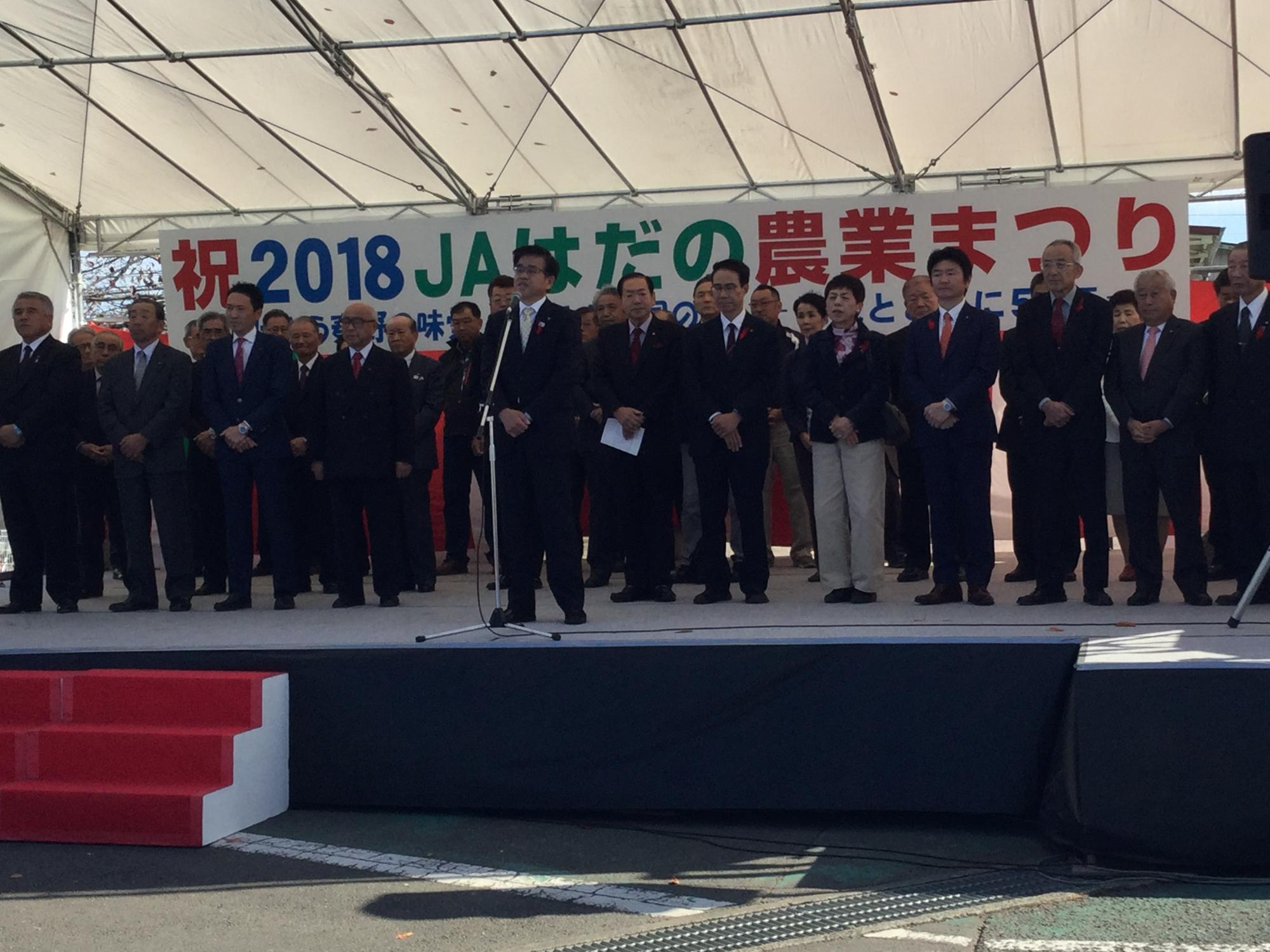 屋外に設置されたステージの上で、スーツ姿の市長が中央のマイクの前で演説をしており、その周囲に多数の男女が立って並び、後方には「祝 2018 JAはだの 農業まつり」と書かれた横断幕が掲げられている写真