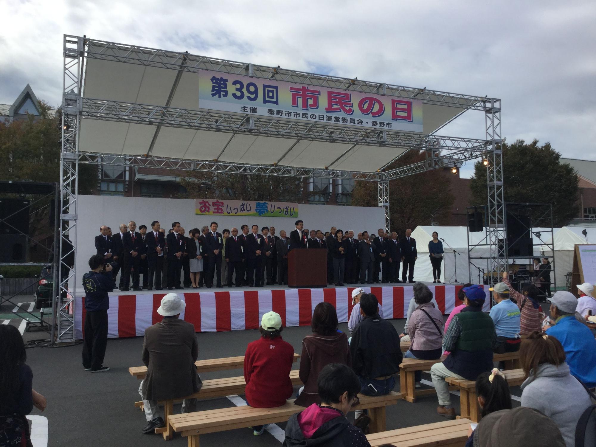 屋外イベント会場の白いテント屋根付きの特設ステージで、スーツ姿の多数の男性が並び、その前に立つ市長を、横断幕の下の「お宝いっぱい夢いっぱい」の垂れ幕と紅白の幕を背景に、木製のベンチに座った多くの観客が見ている市民祭の写真