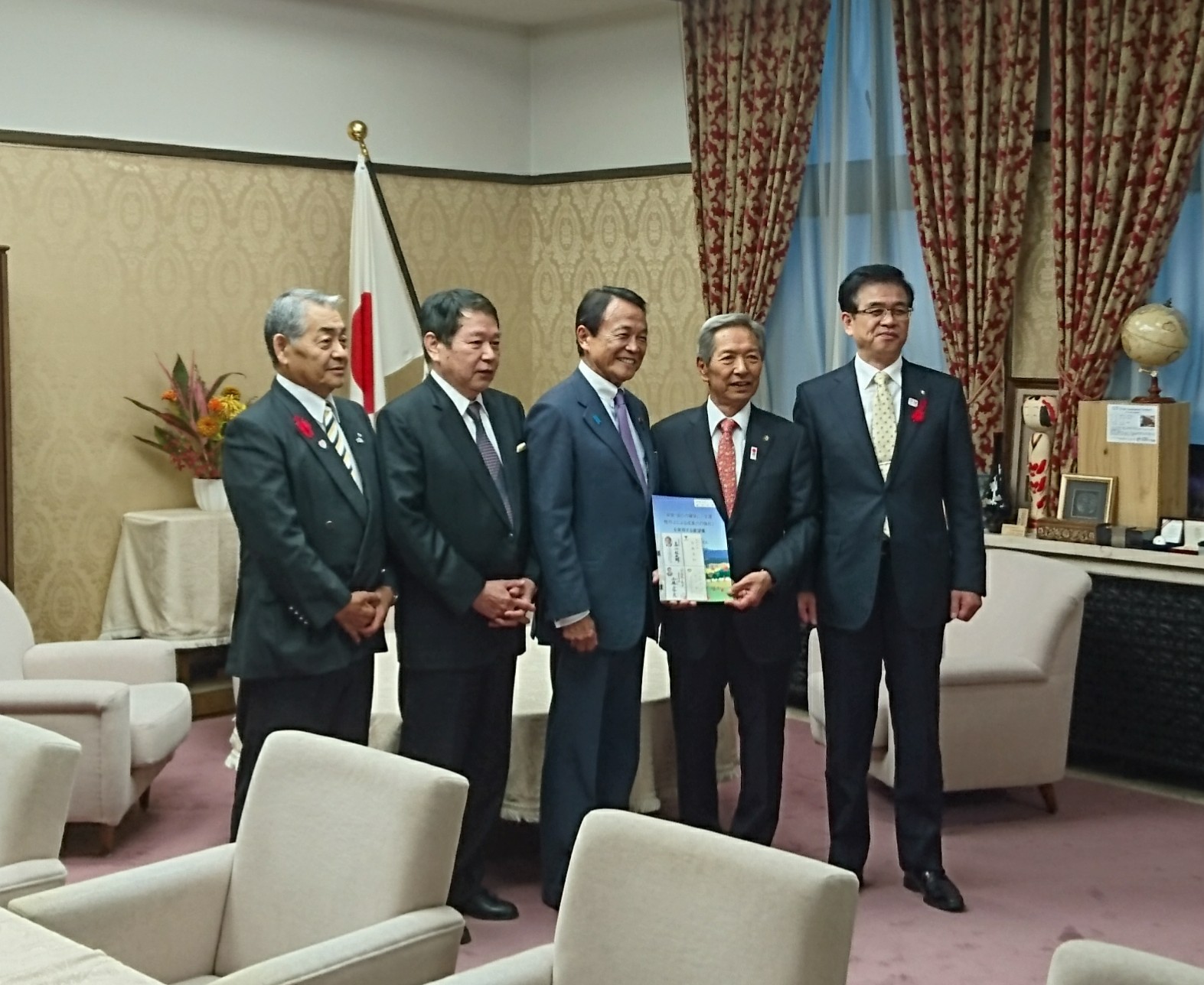 スーツ姿の市長と男性4人が、中央の男性が両手で持っている冊子に注目しながら、日本の国旗の前で直立して並んでいる記念写真